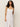 A person with curly hair wears a sleeveless, button-down dress named "wilder linen dress bleached white" by Sanctuary Clothing. They stand against a plain background with a neutral expression, one arm relaxed and the other slightly bent.