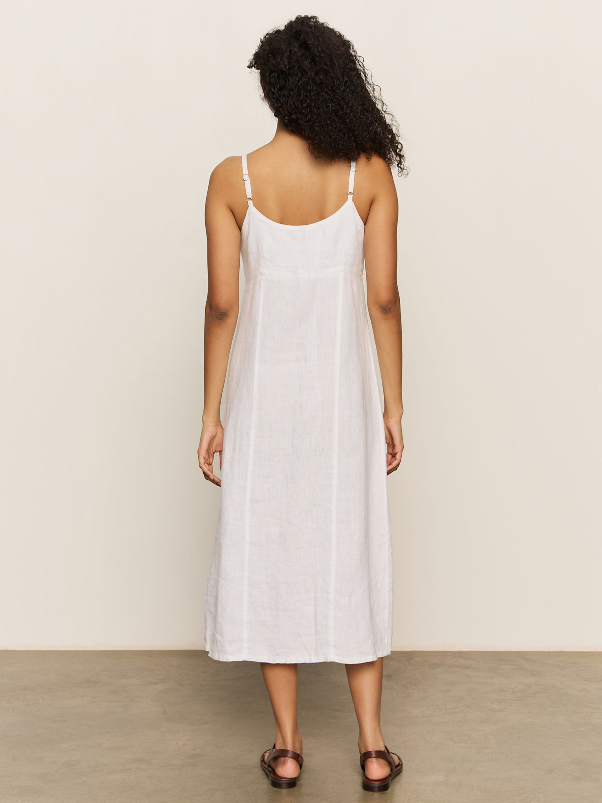 Rear view of a curly-haired person in a Sanctuary Clothing wilder linen dress bleached white and brown sandals, standing on a gray floor against an off-white background.