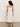 Rear view of a curly-haired person in a Sanctuary Clothing wilder linen dress bleached white and brown sandals, standing on a gray floor against an off-white background.
