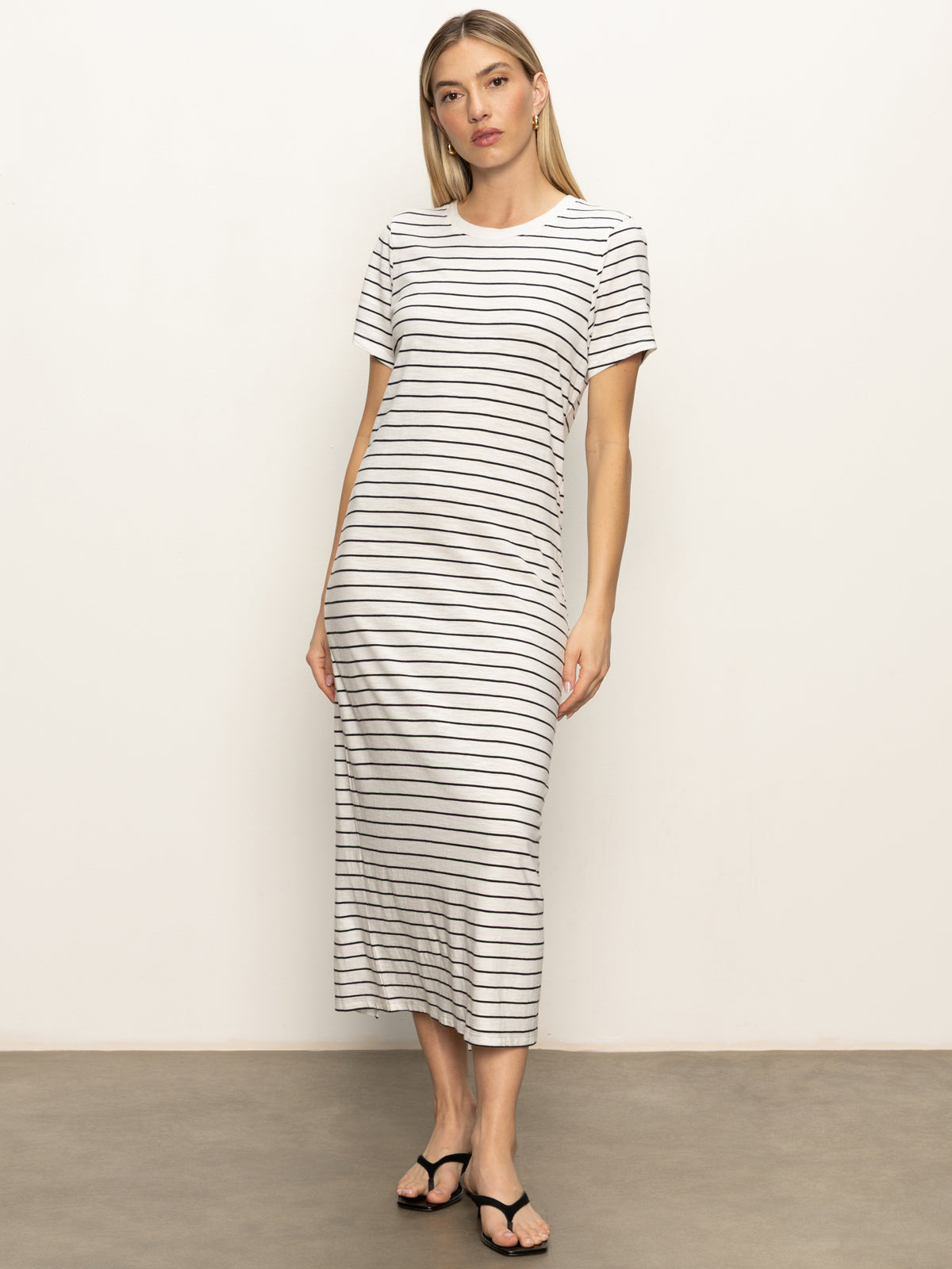 A woman with straight, shoulder-length blonde hair and a neutral expression wears the Sanctuary Clothing perfect tee maxi dress in chalk black stripe and black sandals, standing against a plain background.