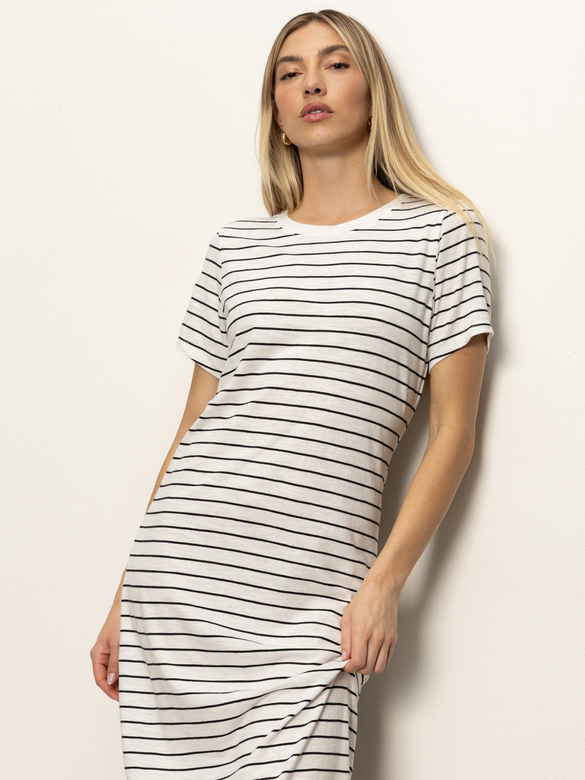 A woman with long blonde hair wears the Sanctuary Clothing perfect tee maxi dress in chalk black stripe, featuring short sleeves and thin black horizontal stripes, standing against a plain light background.