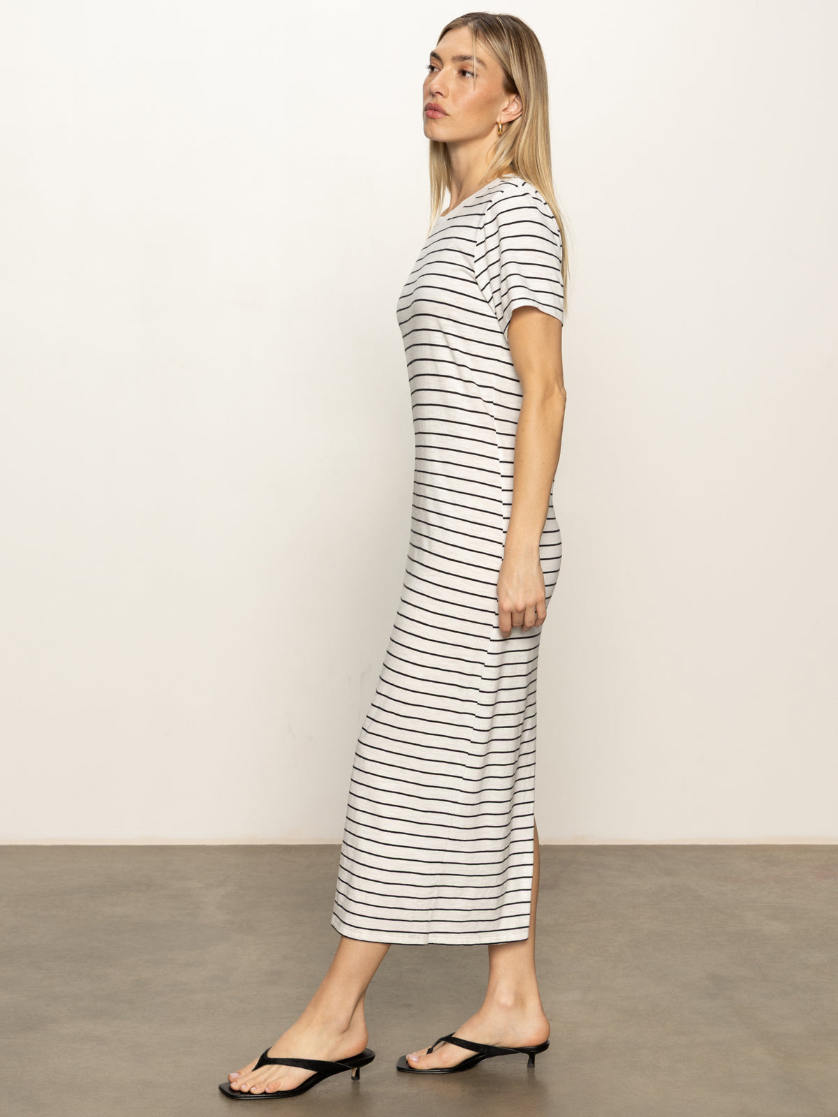 A person with long blonde hair stands in profile against a plain light background, wearing the Sanctuary Clothing perfect tee maxi dress in chalk black stripe paired with black sandals.