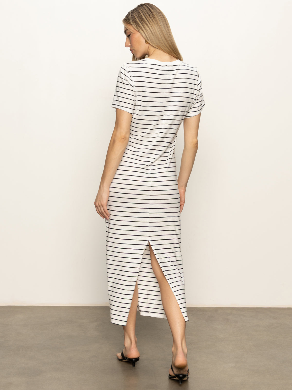 A woman with long blonde hair faces away, wearing the Sanctuary Clothing perfect tee maxi dress in chalk black stripe and black heeled sandals. The dress features short sleeves, black horizontal stripes, and a back slit. Background is plain and light-colored.