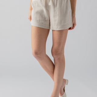 A person is shown from the waist down against a plain background. They are wearing the Always Linen Short Vineyard Stripe by Sanctuary Clothing, featuring beige stripes, a high-waisted fit with a drawstring tie, and a white waistband. Their feet are in white, open-toe sandals with a low heel.