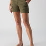 A person is standing against a plain background wearing the Rebel Short Hiker Green from Sanctuary Clothing. These green cargo shorts feature pockets and double buttons. They pair it with a pair of brown sandals. Their right hand is in their pocket while their left hand is relaxed by their side.