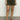 A person wearing a white top and Sanctuary Clothing's "Renegade Short Kalamata" stands on a gray floor, sporting brown buckle-design sandals against a plain white wall.