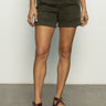 A person wearing a white top and Sanctuary Clothing's "Renegade Short Kalamata" stands on a gray floor, sporting brown buckle-design sandals against a plain white wall.