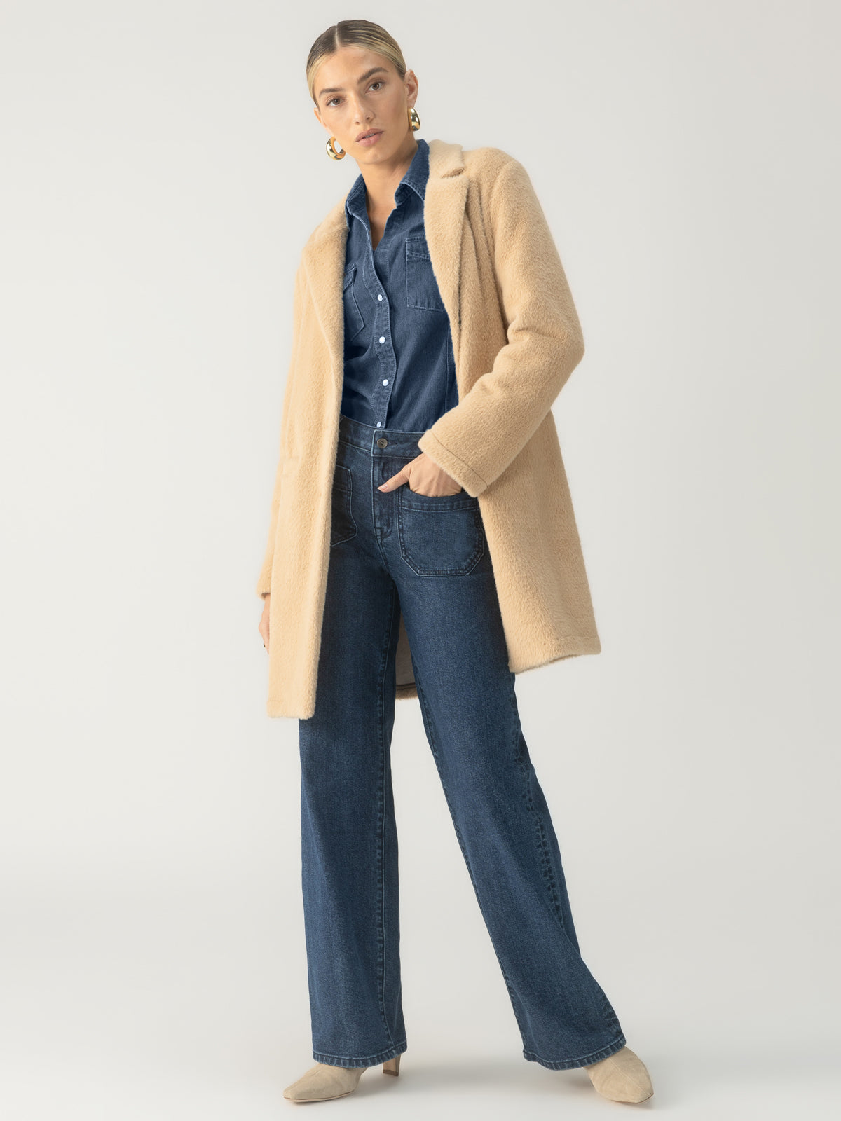 A person with light hair is standing against a plain background. They are wearing the Sanctuary Clothing Hometown Jacket in Camel over a denim shirt and high-waisted jeans. They have one hand in a pocket and are wearing beige shoes. The person has a confident stance and large hoop earrings.