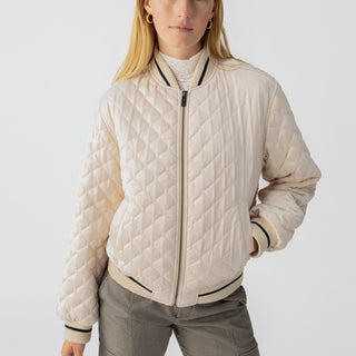 A person with long blonde hair is wearing the Marilyn Bomber Toasted Almond by Sanctuary Clothing, featuring a beige quilted design with a zipper and black striped trim on the collar, cuffs, and hem. They have one hand in their checkered pants pocket and are standing against a plain white background.