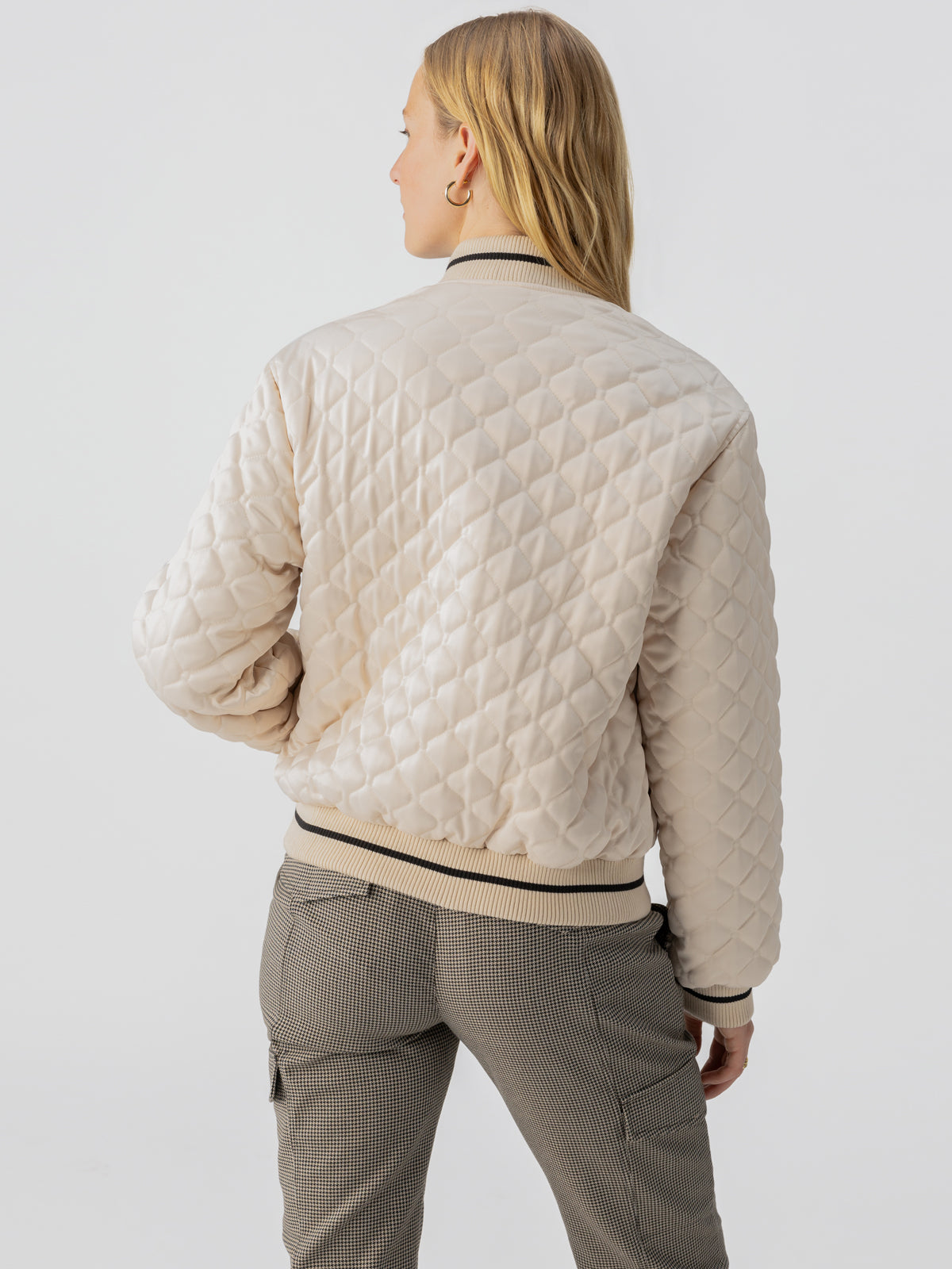 A person with long, blonde hair is seen from the back wearing the Marilyn Bomber Toasted Almond by Sanctuary Clothing, a light beige, quilted bomber jacket featuring a diamond pattern and black and beige striped cuffs and collar. They are also wearing checked pants with side pockets.