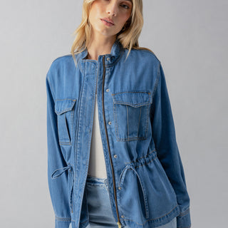 A person with light hair poses against a neutral background, donning the Sanctuary Clothing Cinched Surplus Jacket in Sun Drenched. The blue denim jacket features a front zipper, multiple pockets, and a drawstring waist, worn over a white top. They are looking slightly to the right with a relaxed expression.