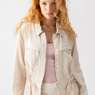 A woman with long, wavy red hair is wearing the Lora Surplus Jacket Crystal Stone by Sanctuary Clothing over a pale pink top. The light beige utility jacket features multiple pockets and drawstring details at the waist. She is also wearing matching light beige pants against a plain white background.