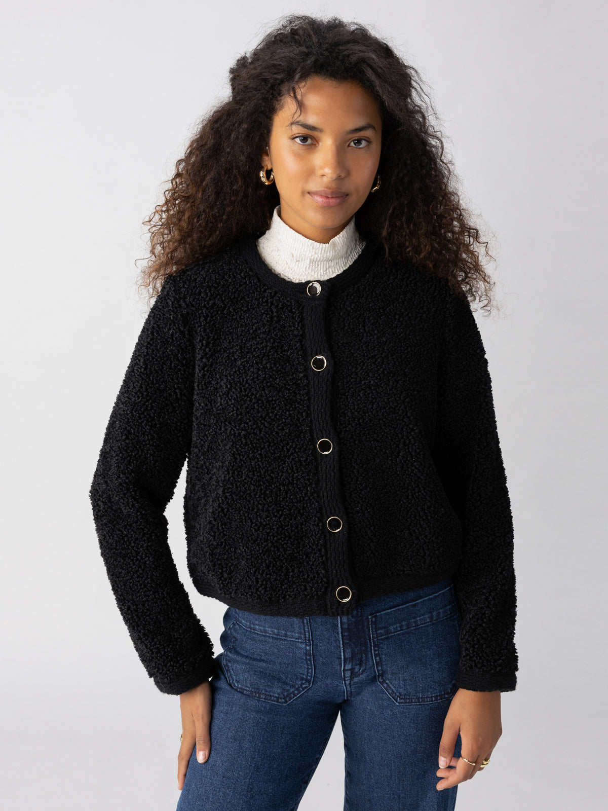 A person with long, curly hair is wearing the Cozy Cardigan Black by Sanctuary Clothing over a white turtleneck and blue jeans. They are standing in front of a plain, light-colored background, looking directly at the camera with a slight smile.