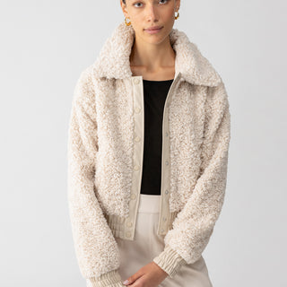 A person with pulled-back hair is wearing the Libby Sherpa Jacket in Toasted Almond by Sanctuary Clothing over a black top. The cream-colored, textured jacket features a high collar, snap buttons, and ribbed cuffs. They are standing against a plain, light-colored background and looking directly at the camera with a neutral expression.