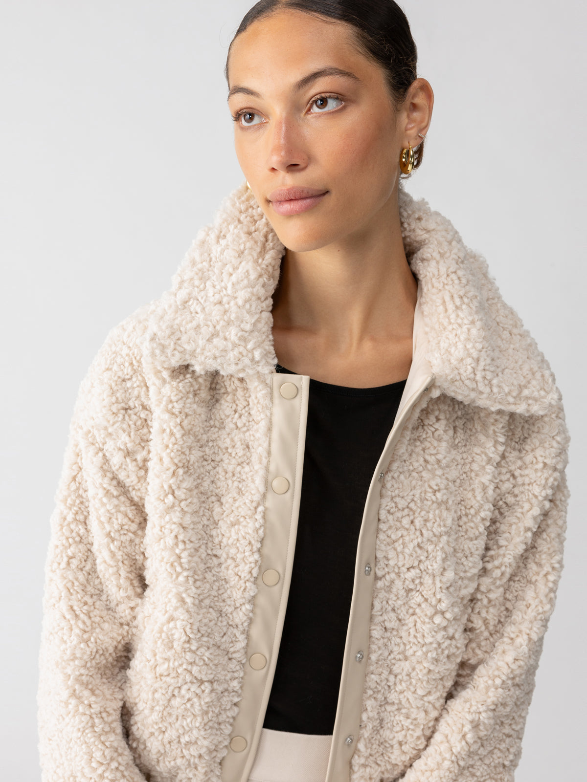 A person with a serene expression looks into the distance. They have their dark hair tied back and are wearing a cozy Libby Sherpa Jacket in Toasted Almond from Sanctuary Clothing over a black top. The jacket features snap buttons and a large collar, and they are wearing gold earrings.