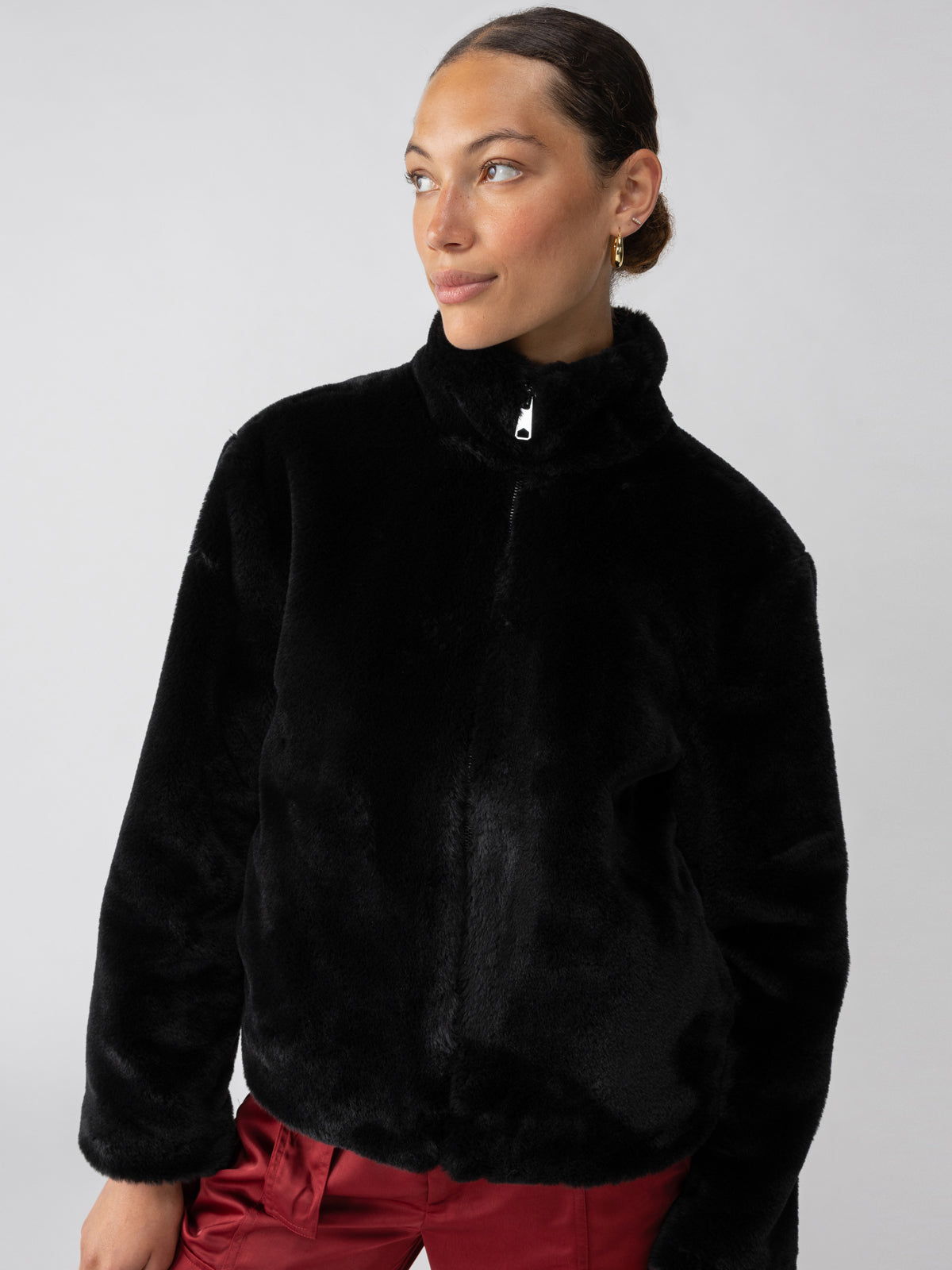 A person wearing the Lux Fur Coat Black by Sanctuary Clothing and red pants poses against a plain background, gazing to the right. The coat features a high collar, and the person has neatly pulled-back hair and is wearing hoop earrings.