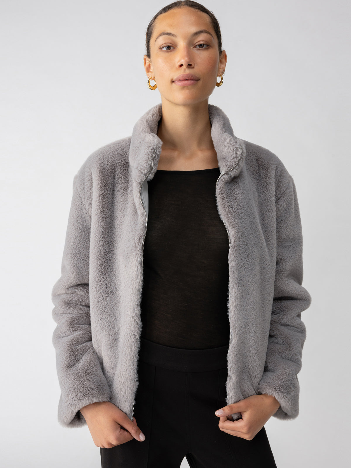 A person with tied-back hair and gold hoop earrings is wearing a Lux Fur Coat Silver Fox by Sanctuary Clothing over a black top, standing against a plain light background. They have a composed expression and their hands in the pockets of the coat.