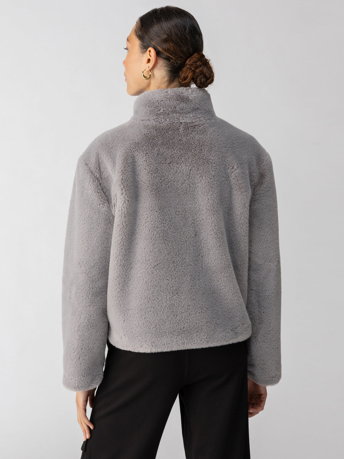 A person with their back to the camera is wearing Sanctuary Clothing's Lux Fur Coat Silver Fox and black pants. Their hair is tied back in a low bun, and they have a single gold hoop earring in their left ear. The background is plain and light gray.