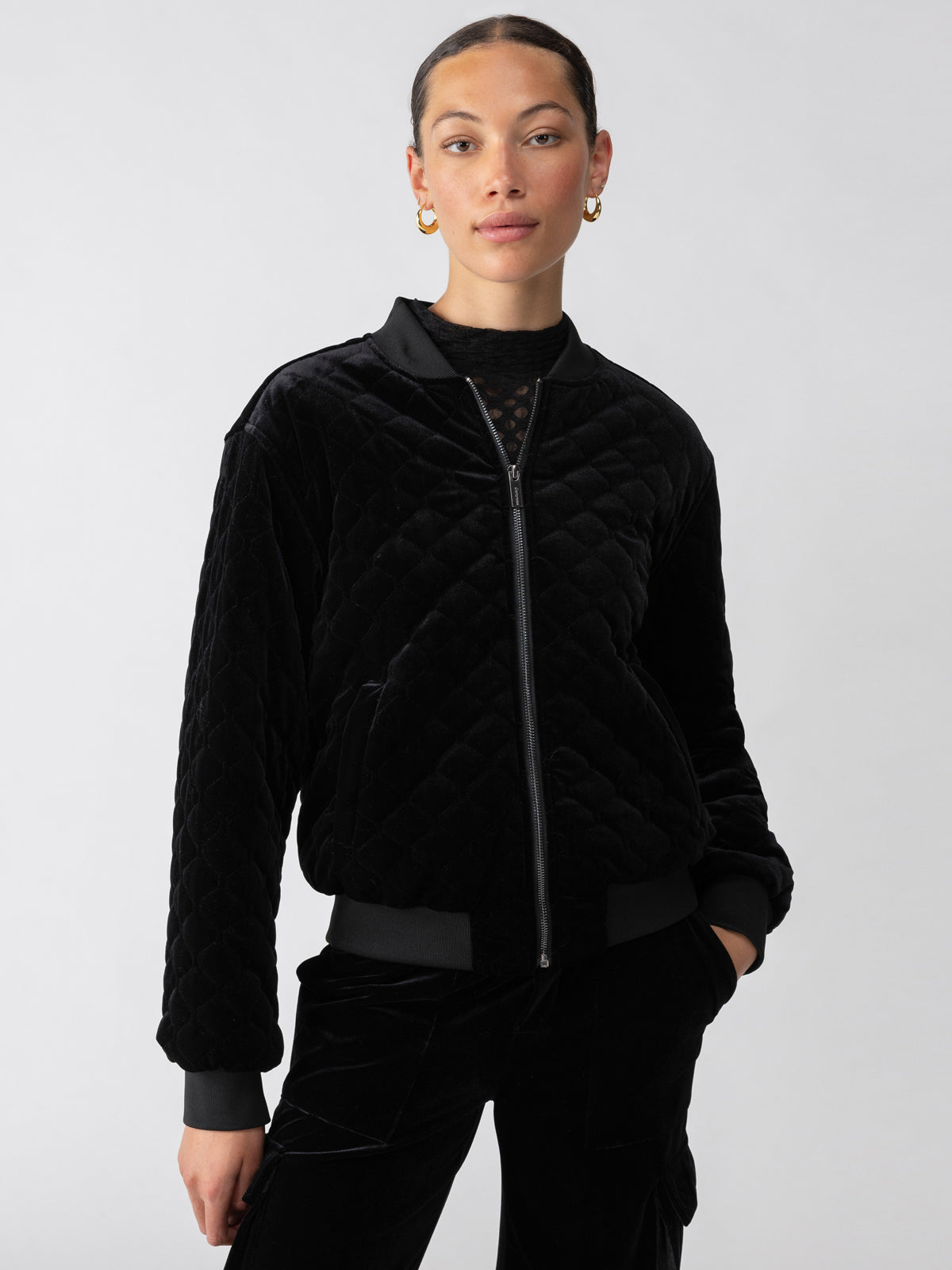 A person stands against a plain background wearing the Mona Velvet Bomber Black by Sanctuary Clothing. The jacket features a zip-up front and ribbed cuffs and collar. They have short hair pulled back, are wearing gold hoop earrings, and have a confident expression. Their hands are relaxed in the jacket pockets.