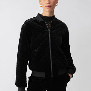 A person stands against a plain background wearing the Mona Velvet Bomber Black by Sanctuary Clothing. The jacket features a zip-up front and ribbed cuffs and collar. They have short hair pulled back, are wearing gold hoop earrings, and have a confident expression. Their hands are relaxed in the jacket pockets.