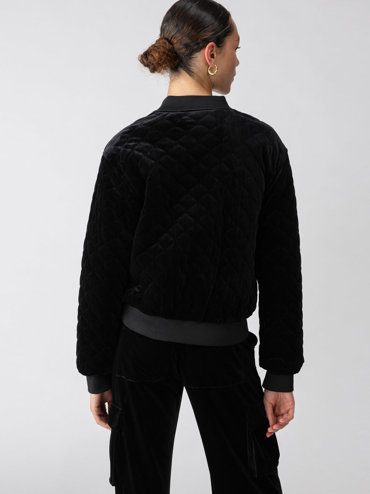 The image shows a person with neatly tied-back hair from the back, wearing the Mona Velvet Bomber Black by Sanctuary Clothing paired with black pants. The background is plain gray.