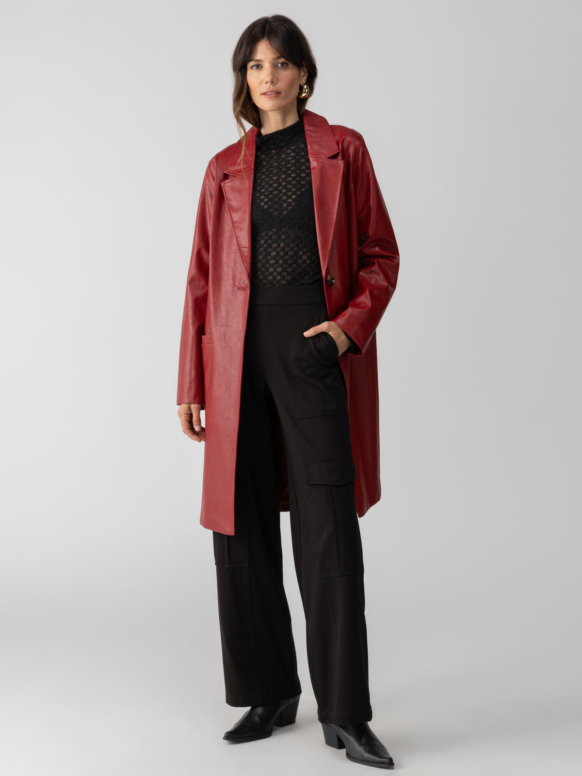 A woman stands against a plain background wearing the Sanctuary Clothing Vegan Leather Coat Garnet over a black, semi-sheer, patterned top and black wide-leg pants with pockets. She completes her look with black-heeled shoes.