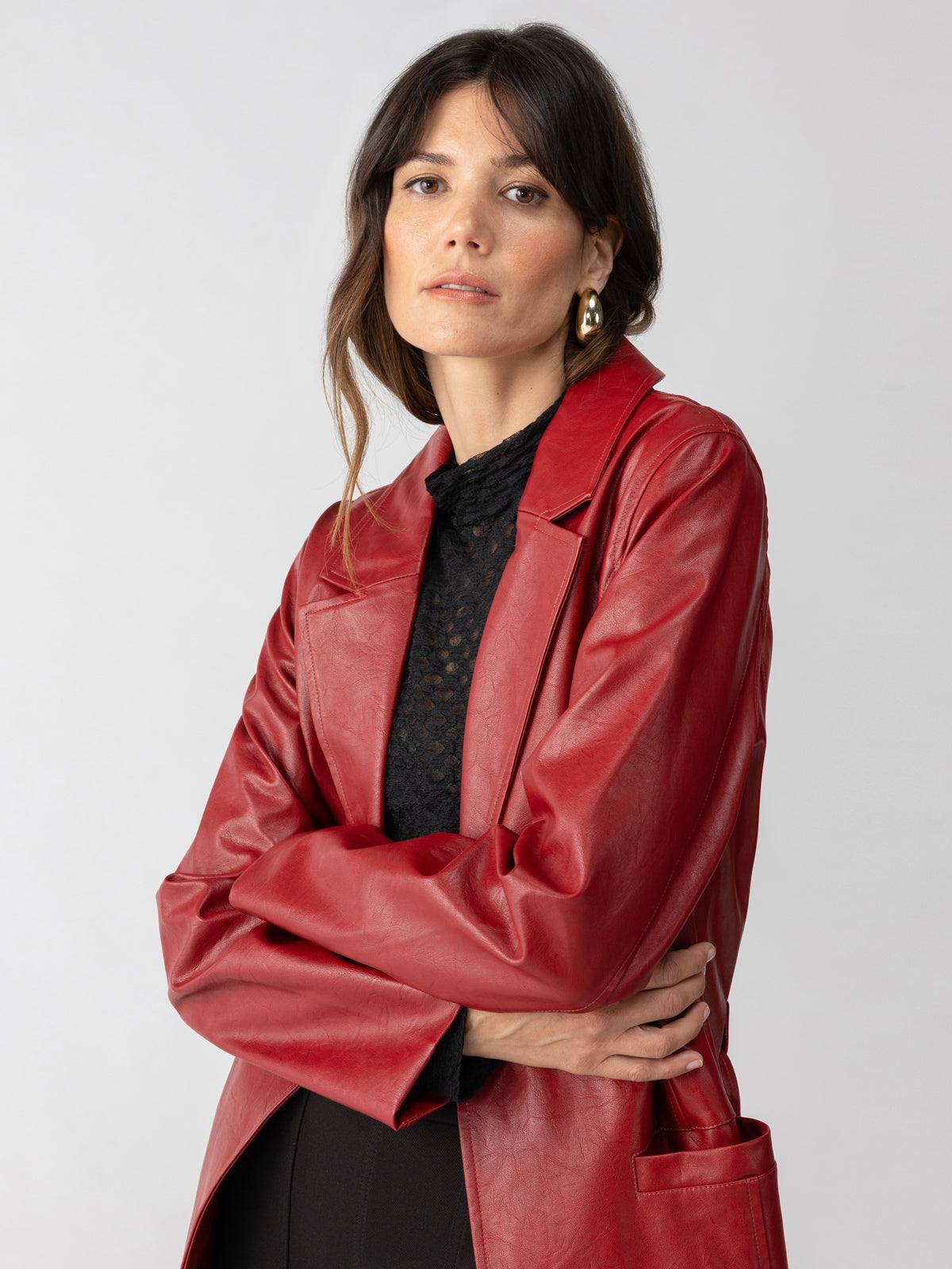 A woman with long brown hair is wearing the Vegan Leather Coat Garnet by Sanctuary Clothing over a black top. She has gold hoop earrings and is standing against a plain grey background, with her arms crossed and a neutral expression on her face.