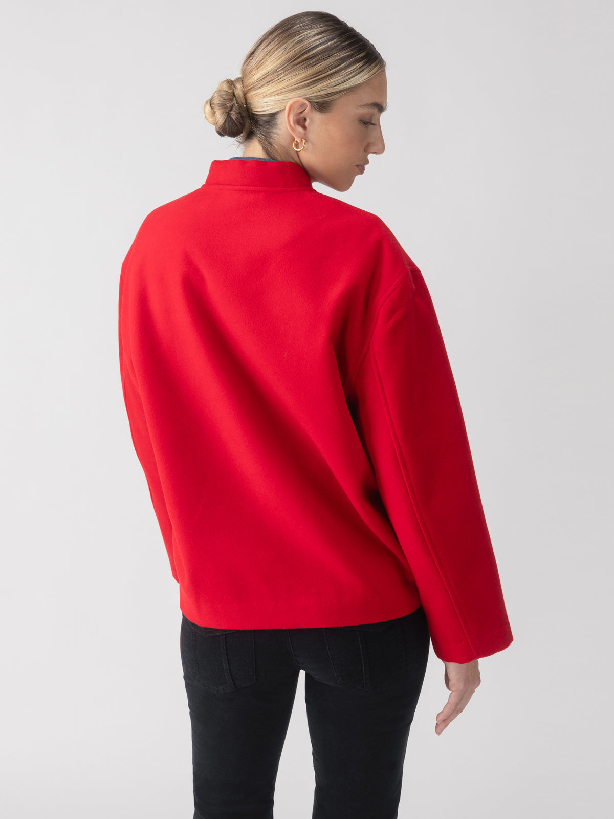 A person with light hair styled in a low bun is wearing the Modern Bomber Jacket Mars Red from Sanctuary Clothing and black pants, standing with their back turned and head slightly turned to the side against a plain white background.