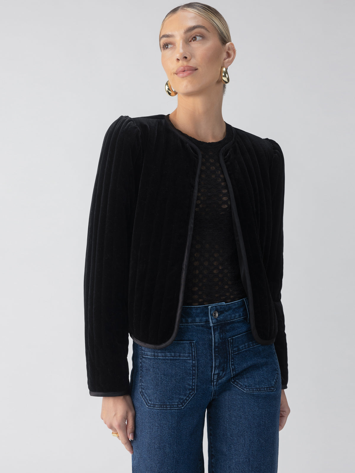 A person wearing the Sanctuary Clothing Quilted Velvet Jacket in black over a sheer black top and high-waisted blue jeans. They have blonde hair pulled back and are accessorized with gold hoop earrings, standing against a light gray background.