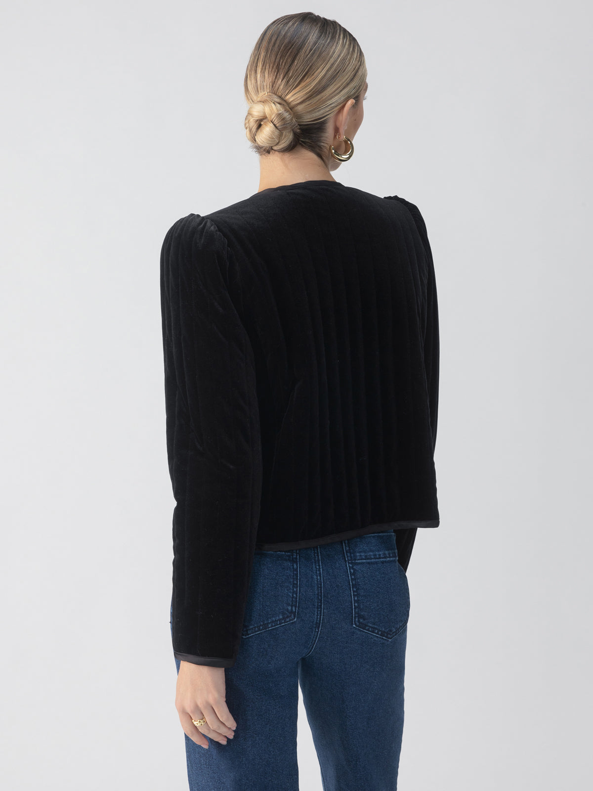 A person with blonde hair in a bun is wearing a Sanctuary Clothing Quilted Velvet Jacket in black paired with blue jeans. They are standing with their back to the camera against a plain background.