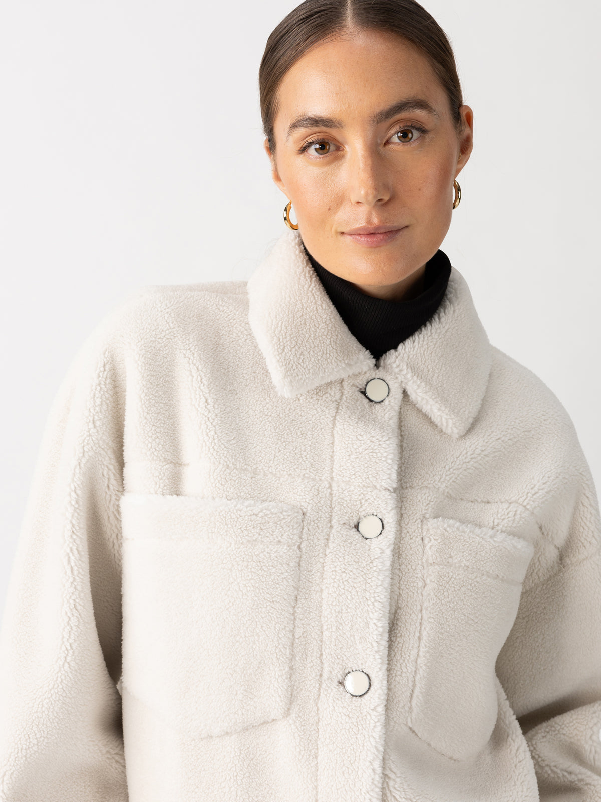 A person is wearing the "Cozy Sherpa Shacket Chalk" by Sanctuary Clothing, layered over a black turtleneck. Their hair is neatly tied back, and they are accessorized with gold hoop earrings against a plain white background.