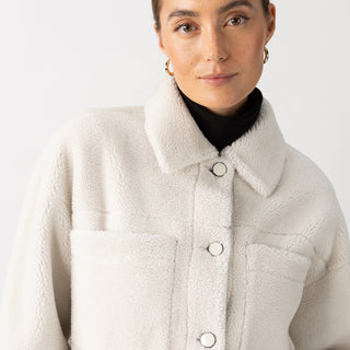 A person is wearing the "Cozy Sherpa Shacket Chalk" by Sanctuary Clothing, layered over a black turtleneck. Their hair is neatly tied back, and they are accessorized with gold hoop earrings against a plain white background.