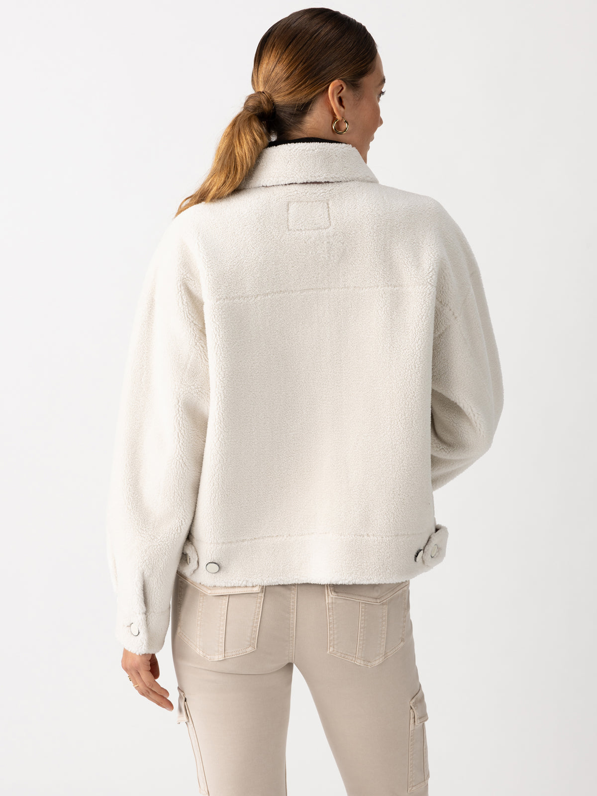 A person with long hair in a ponytail is wearing the Cozy Sherpa Shacket Chalk, a white textured jacket from Sanctuary Clothing featuring snap buttons at the cuffs. They are turned away, displaying the back of the jacket. The person pairs it with light beige pants equipped with pockets.