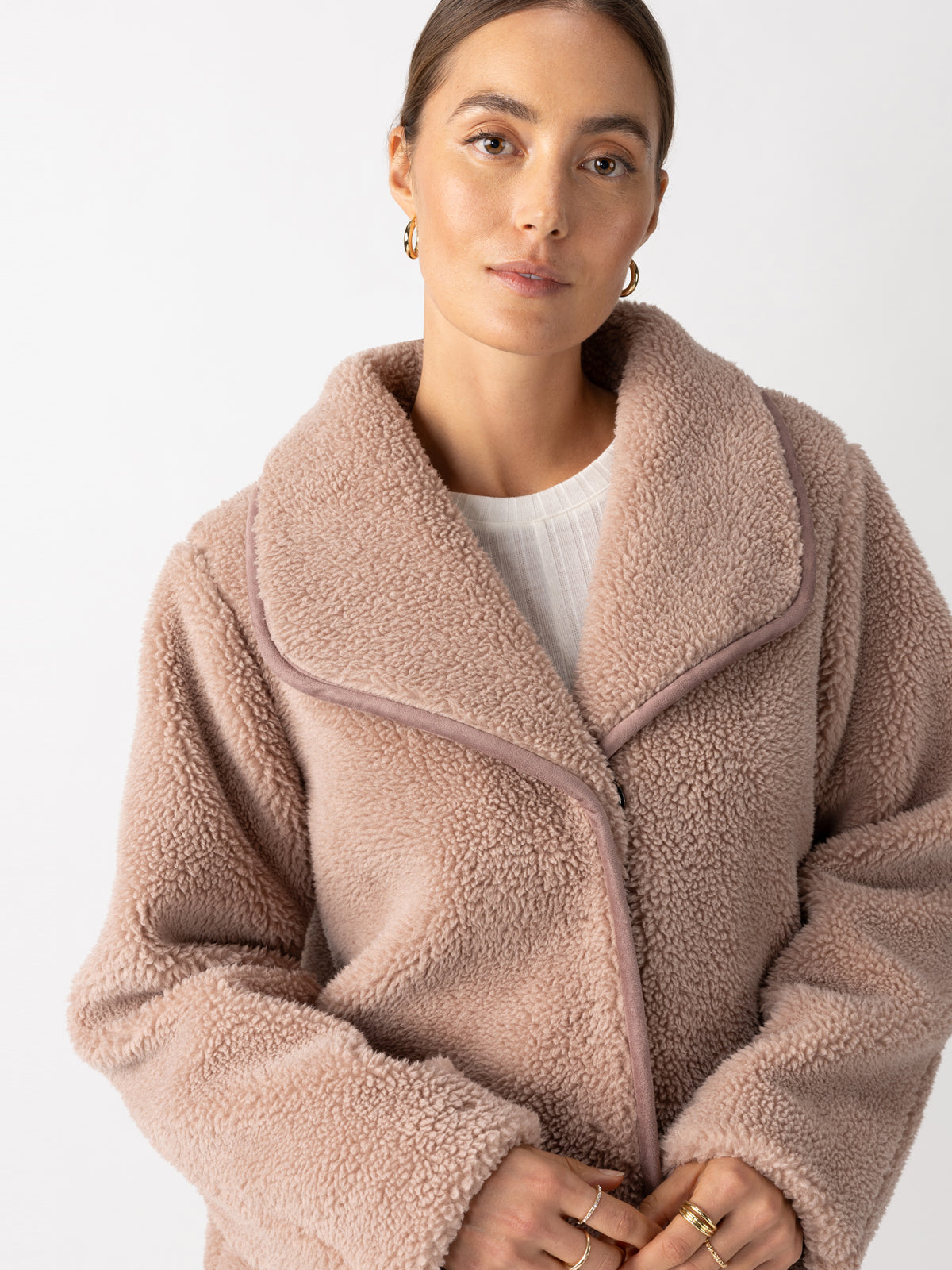 A person wearing the Holly Sherpa Jacket Feather by Sanctuary Clothing, in a cozy, light brown shade with a thick collar, stands against a plain white background. They have long hair pulled back and are accessorized with gold hoop earrings, maintaining a relaxed expression.