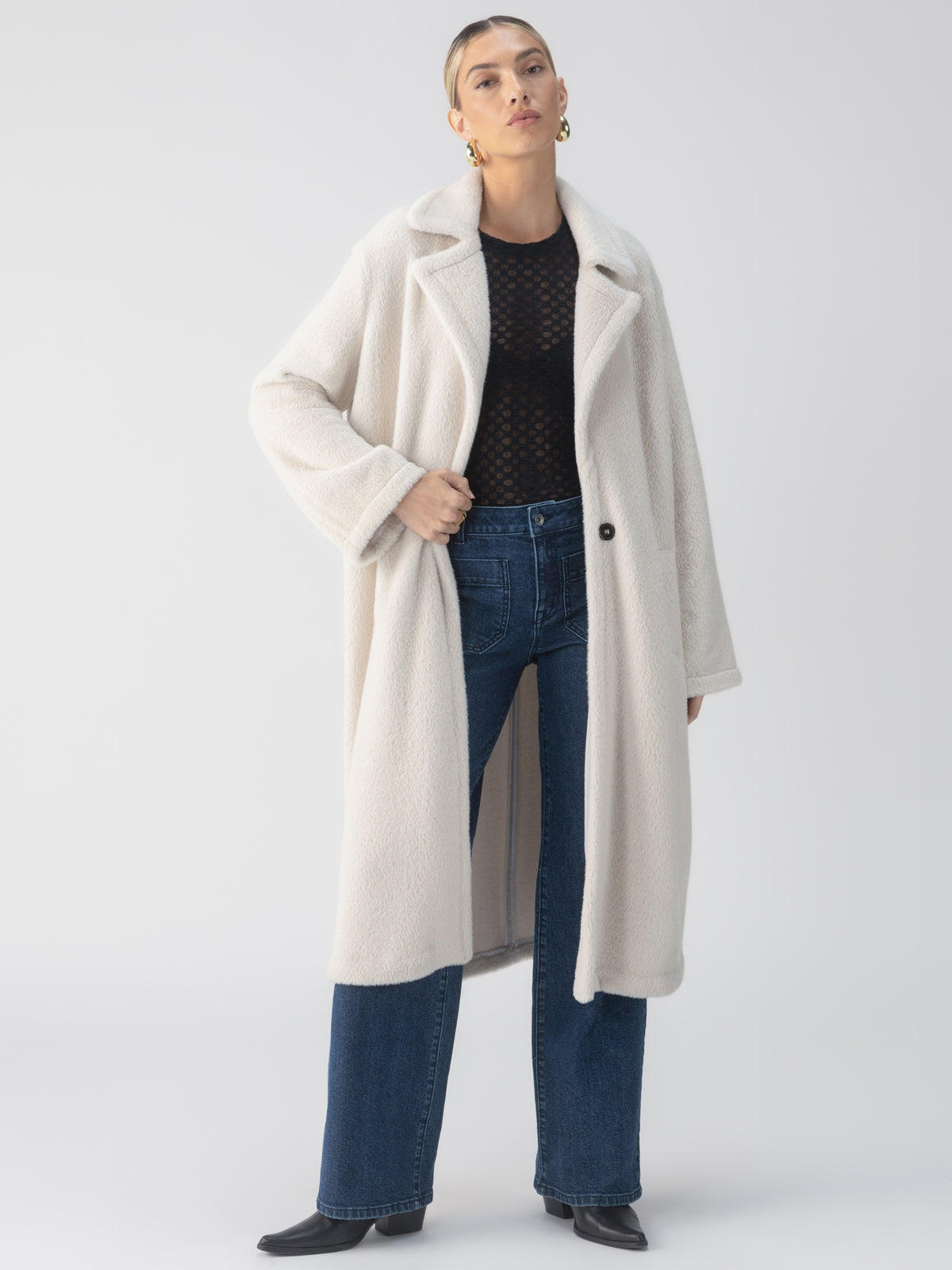 A person stands confidently wearing the "Live In Fur Coat Frosted White" by Sanctuary Clothing over a sheer black top and blue jeans. Their short hair is complemented by large hoop earrings as they strike a fashion-forward pose against a light gray background.