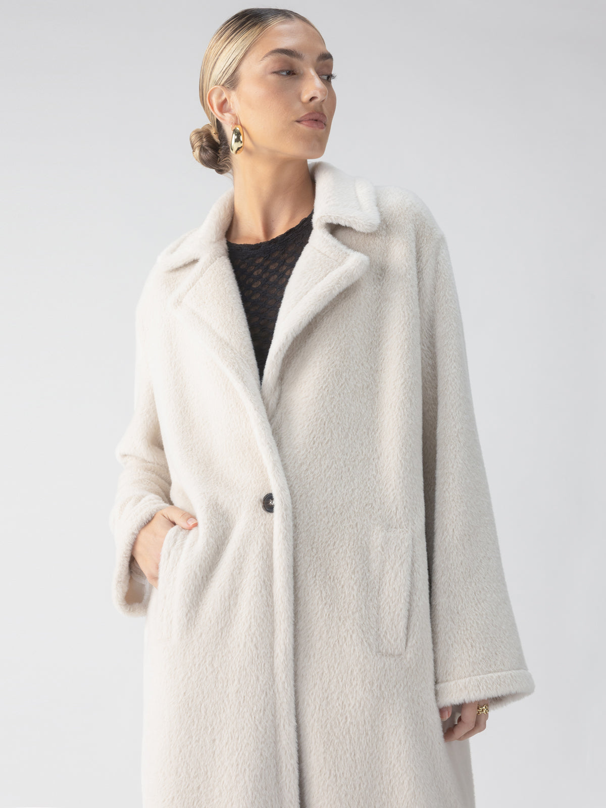 A person wearing the Sanctuary Clothing 'Live In Fur Coat' in Frosted White, featuring a single black button, stands against a light background. Their hair is styled in a low bun, and they accessorize with small hoop earrings. They gaze slightly to the side while keeping one hand in the coat pocket.