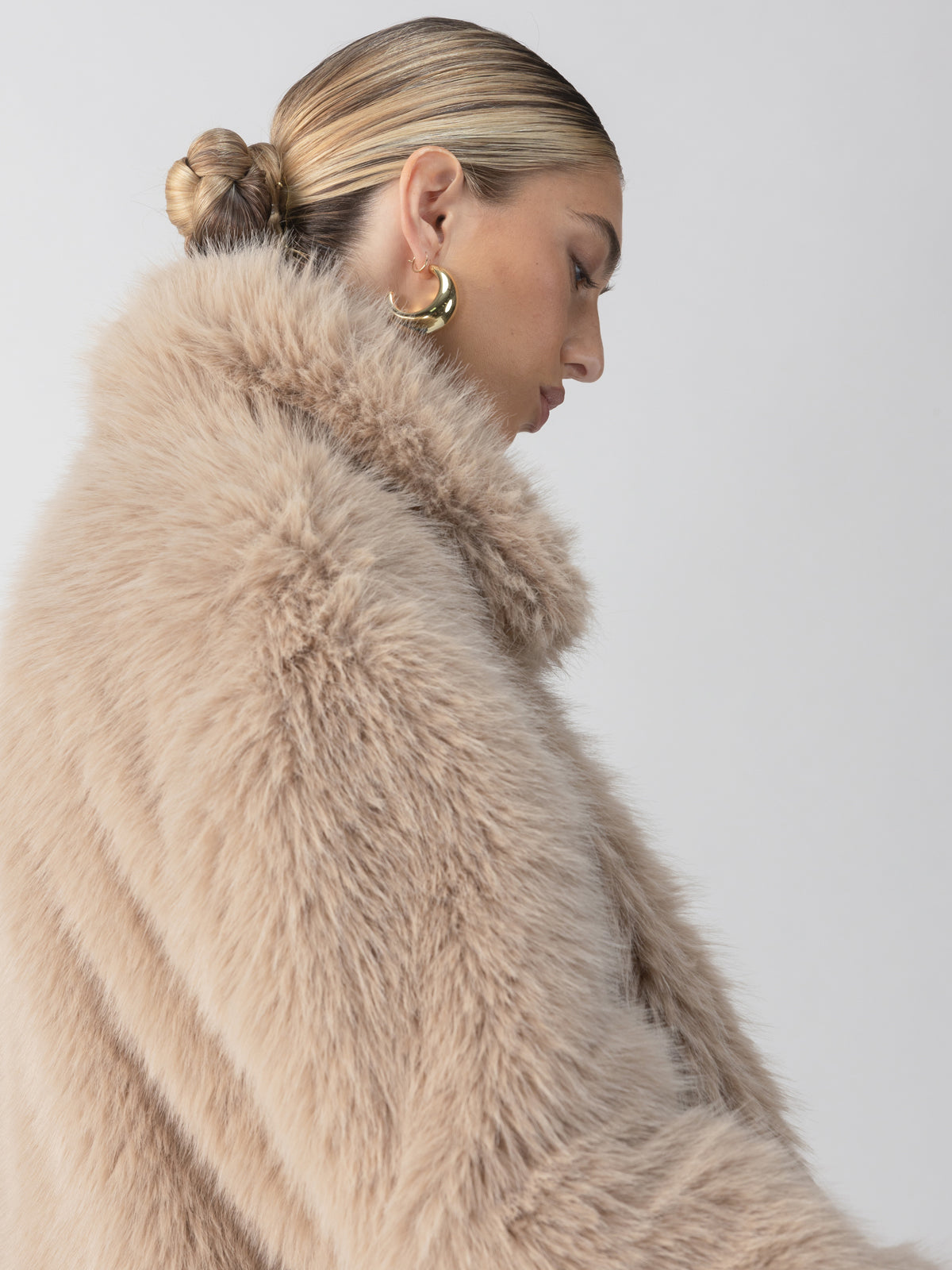 A person with blonde, slicked-back hair styled in a bun is shown in profile. They are wearing the Carmen Fur Coat in Chocolate Sundae by Sanctuary Clothing and gold hoop earrings. The background is plain and white.