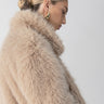 A person with blonde, slicked-back hair styled in a bun is shown in profile. They are wearing the Carmen Fur Coat in Chocolate Sundae by Sanctuary Clothing and gold hoop earrings. The background is plain and white.