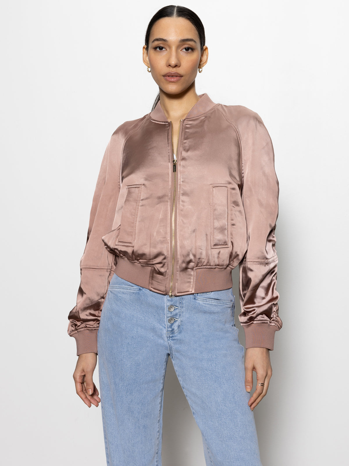 A person wearing a Sanctuary Clothing satin ruched bomber jacket in rose gold and light blue jeans stands against a plain white background. The jacket features a front zipper and ribbed cuffs. The person's hair is pulled back.