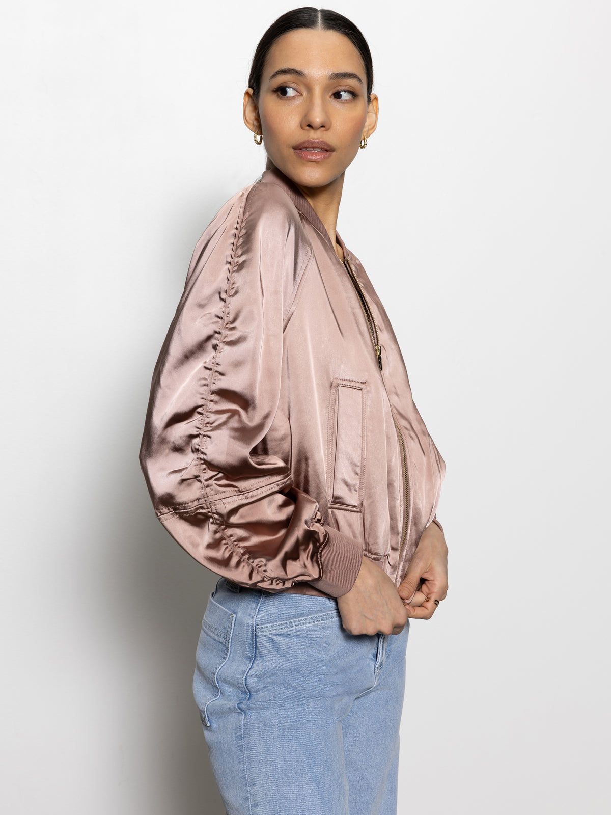 A person with tied-back dark hair wears a satin ruched bomber jacket in rose gold from Sanctuary Clothing, paired with light blue jeans. They are standing against a plain background, looking to the side.