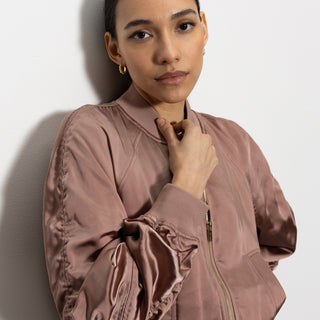 A person with long hair pulled back stands against a wall, wearing a satin ruched bomber jacket in rose gold by Sanctuary Clothing. They are looking at the camera with a calm expression, one hand touching their chest.