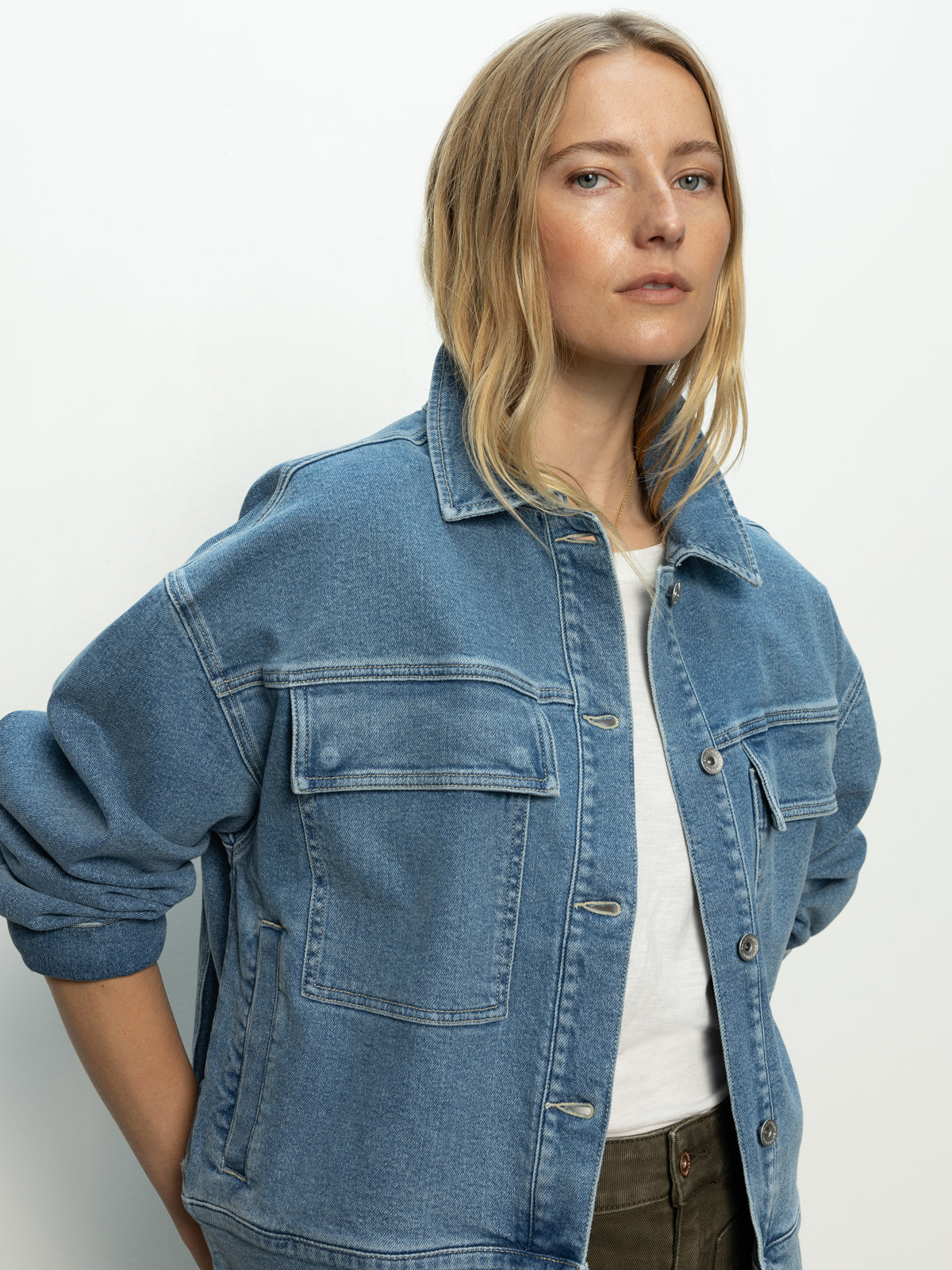 A confident person with long blonde hair, donning Sanctuary Clothing's "Make Way Denim Jacket Wayfarer" with front pockets and rolled-up sleeves, stands against a light background over a white shirt.