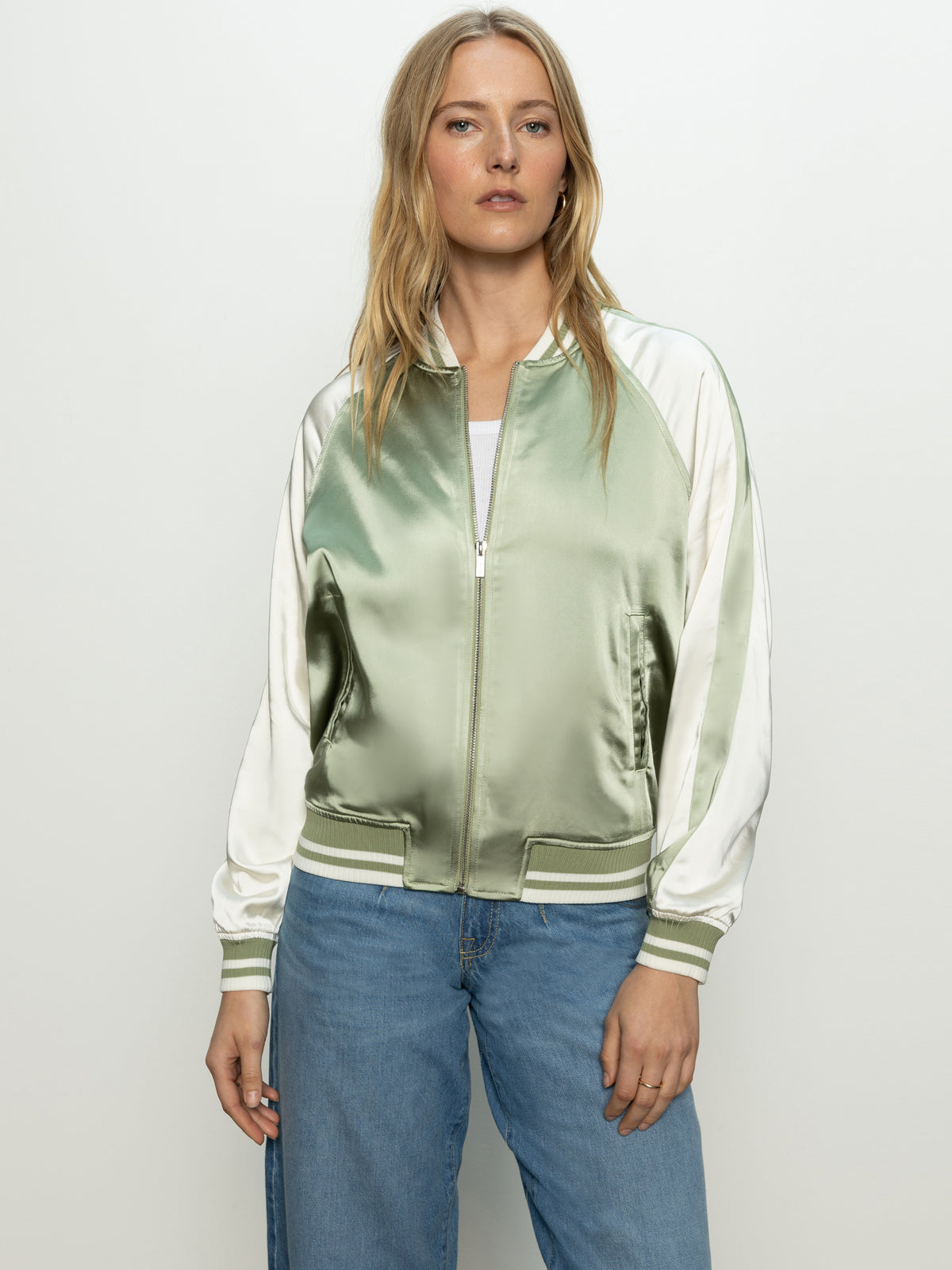 A woman with long blonde hair stands against a neutral background, wearing the Sanctuary Clothing's souvenir jacket in sage—a green and white satin bomber jacket with front zipper and striped cuffs—paired with blue jeans.