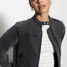 Clad in a Sanctuary Clothing denim moto jacket in dark asphalt, an individual stands against a plain white background. With dark hair pulled back, they wear a white patterned top and maintain a neutral expression while looking at the camera.