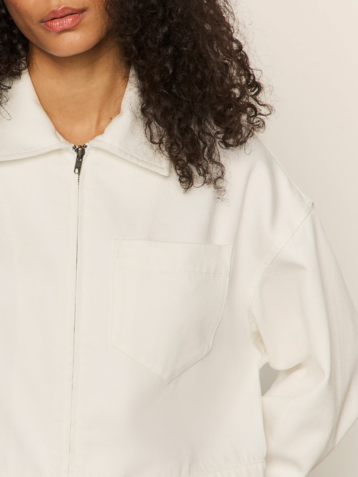 A person with curly dark hair models the Sanctuary Clothing cropped paper boy jacket in bleached white, featuring a collar and chest pocket. The image is cropped at the mouth, omitting the full face and lower part of the jacket.