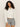 A woman with curly hair models the Sanctuary Clothing cropped paper boy jacket in bleached white, featuring a chest pocket, against a plain beige background.