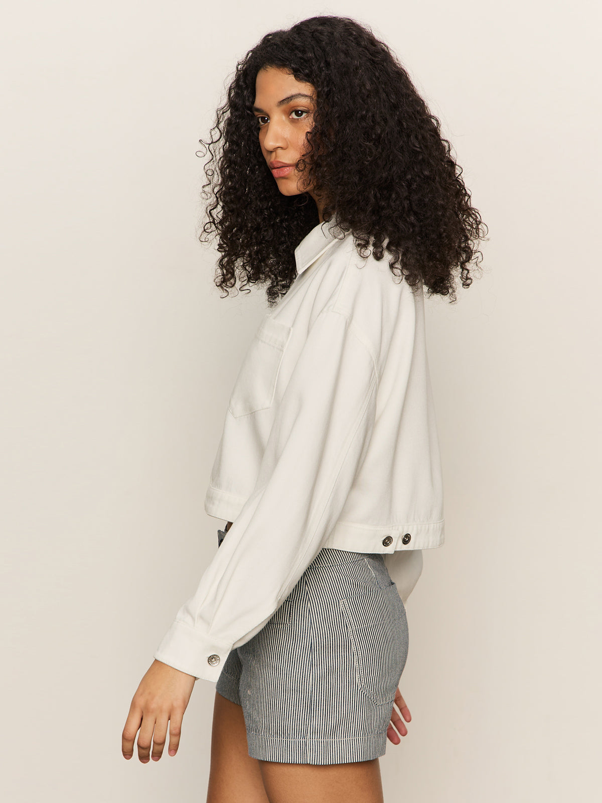A person with curly hair models the Sanctuary Clothing cropped paper boy jacket in bleached white, paired with gray pinstriped shorts, standing sideways against a plain light background with a neutral expression.