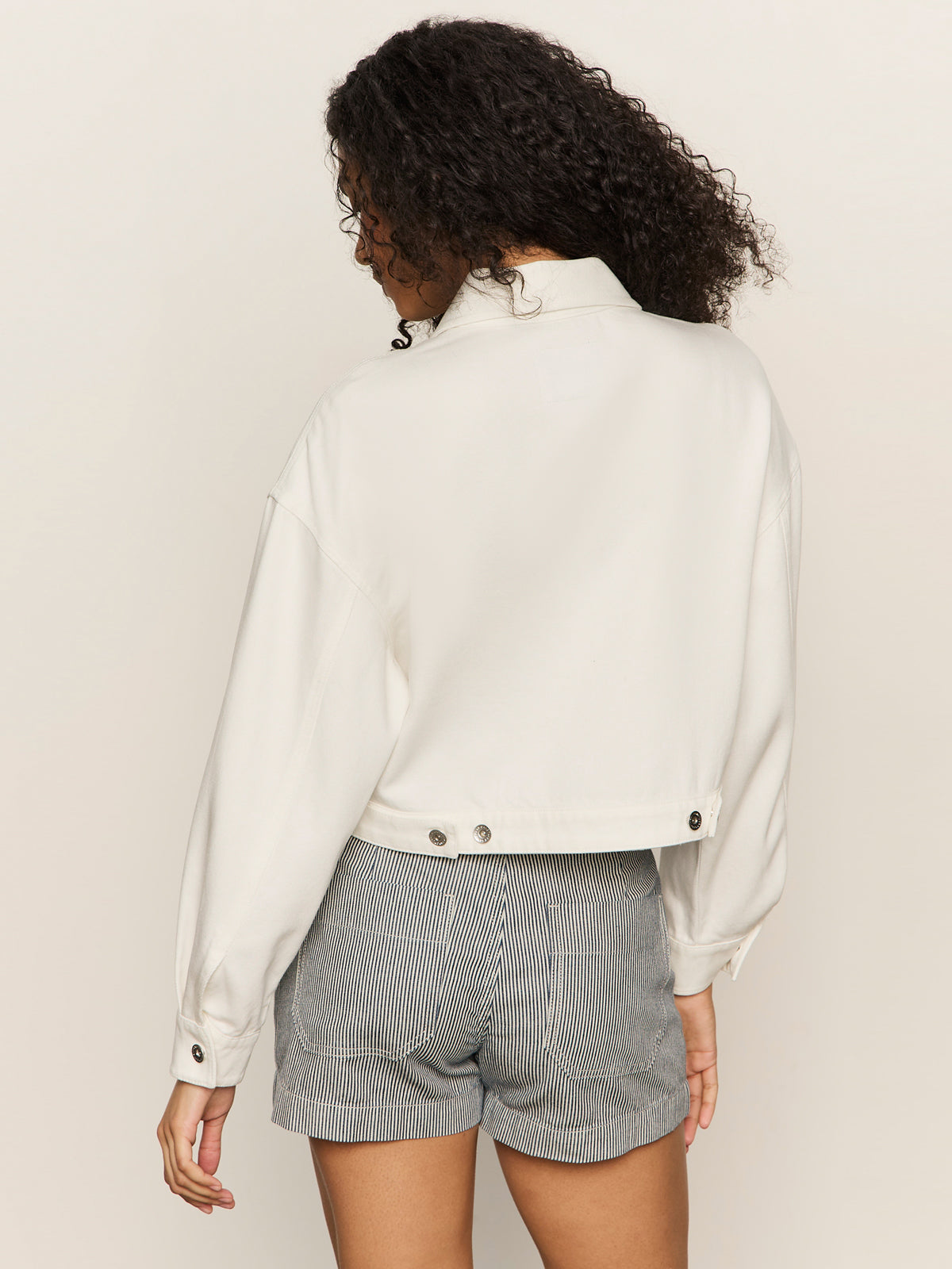 A woman with curly hair is seen from the back, wearing Sanctuary Clothing's cropped paper boy jacket in bleached white, paired with high-waisted, pinstriped grey shorts against a plain light background.