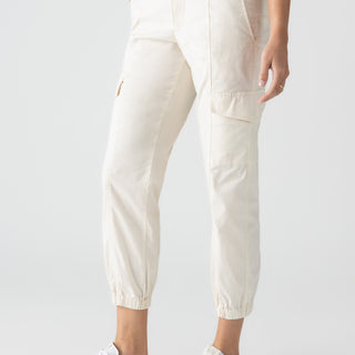 A person wearing the Rebel Pant Eco Natural by Sanctuary Clothing, featuring multiple pockets and cinched cuffs, paired with white sneakers with beige accents. The person is standing against a plain light background with their hands at their sides. The upper body is mostly out of the frame.