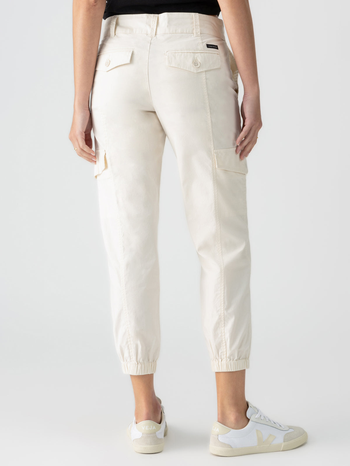 A person is standing with their back facing the camera, wearing the Rebel Pant Eco Natural from Sanctuary Clothing, featuring beige jogger pants with side cargo pockets, buttoned rear pockets, and elastic cuffs at the ankles. They are also wearing white sneakers with a beige sole and 'V' logo details.